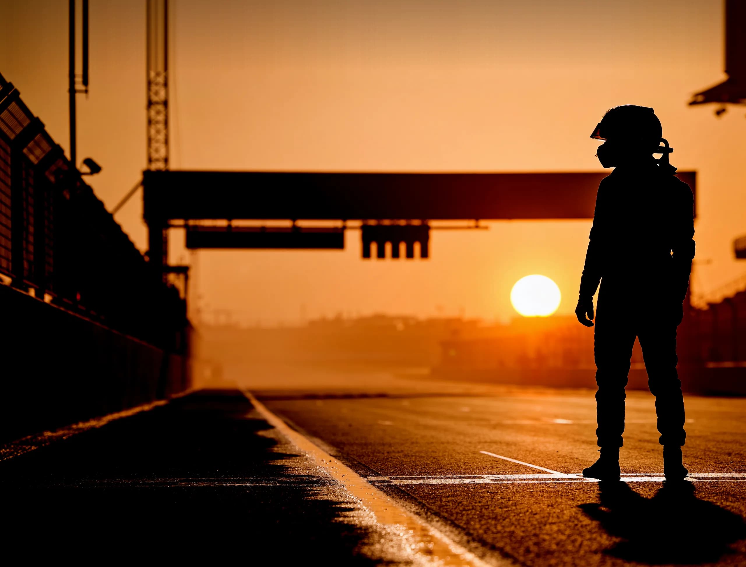 A driver in a plain race suit and full-face helmet, alone in silhouette on a pit straight at sunset, the low sun sitting on the horizon behind them and the wet asphalt running away into haze.